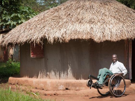 A man smiling sitting on a wheelchair balancing on 2 wheels in a village and