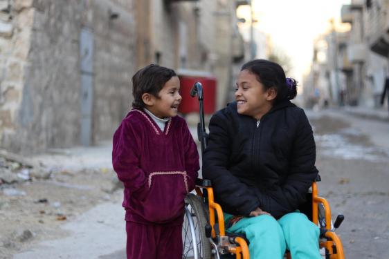 2 children in the streets are looking and smiling at each other, one of whom is using a wheelchair