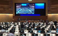 Picture of the podium of room XIX at Palais de Nations, with a big screen on top and crowd